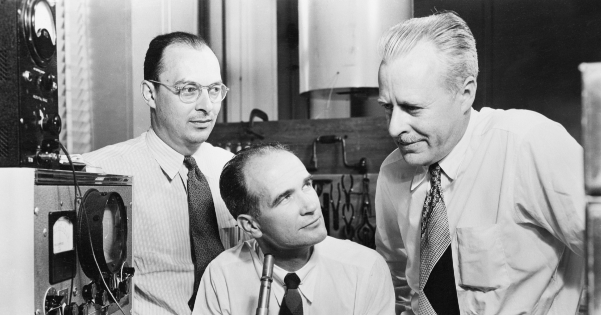 Three men in mid-century attire collaborating around early computing equipment